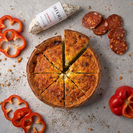 Cheese pizza with slices cut, surrounded by sliced red peppers and salami on a gray surface.